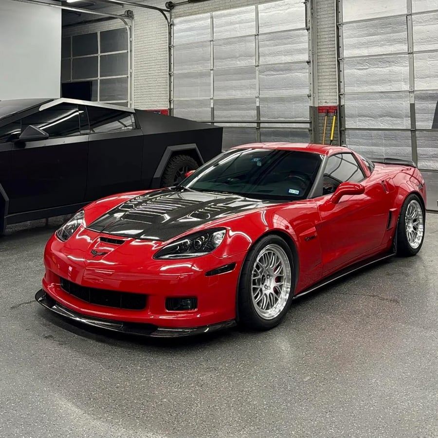 A red sports car with a black hood is parked indoors on a smooth floor, with a futuristic black vehicle visible in the background near large garage doors.