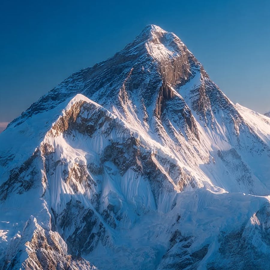 Snow-covered mountain peaks bathed in sunlight under a clear blue sky, with sharp ridges and deep shadows highlighting the rocky, icy landscape.