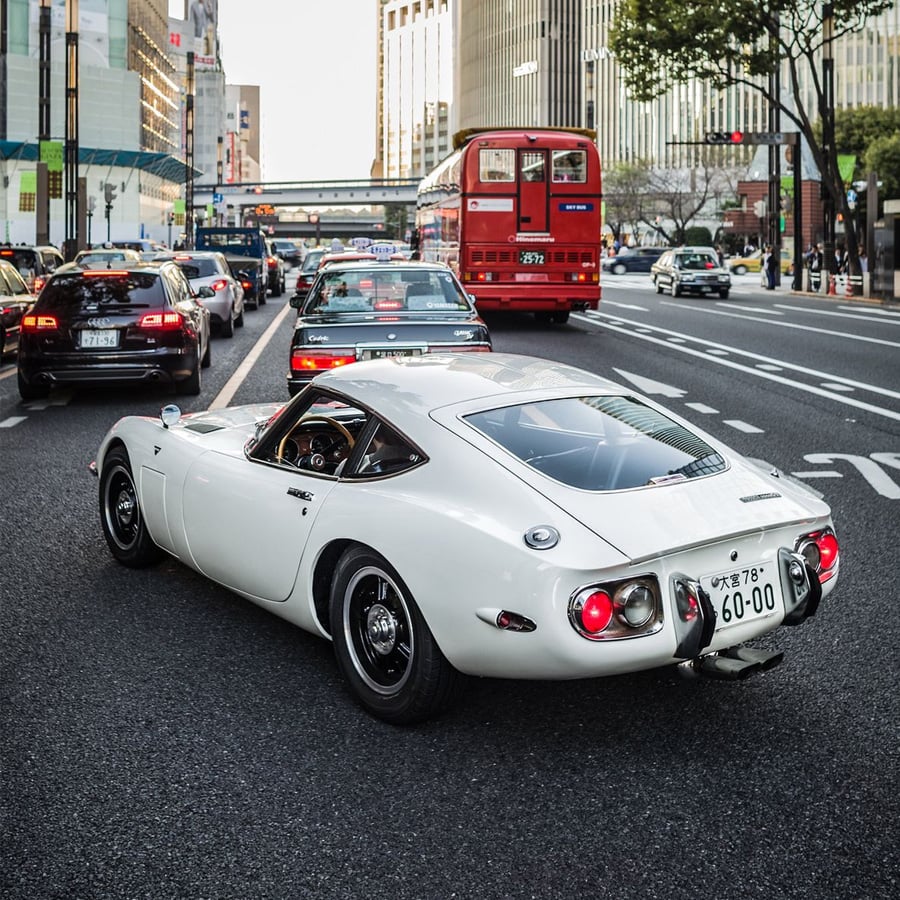 A white classic sports car drives on a city street amid modern cars and taxis. Tall buildings and a red bus are in the background, with trees lining the sidewalk.