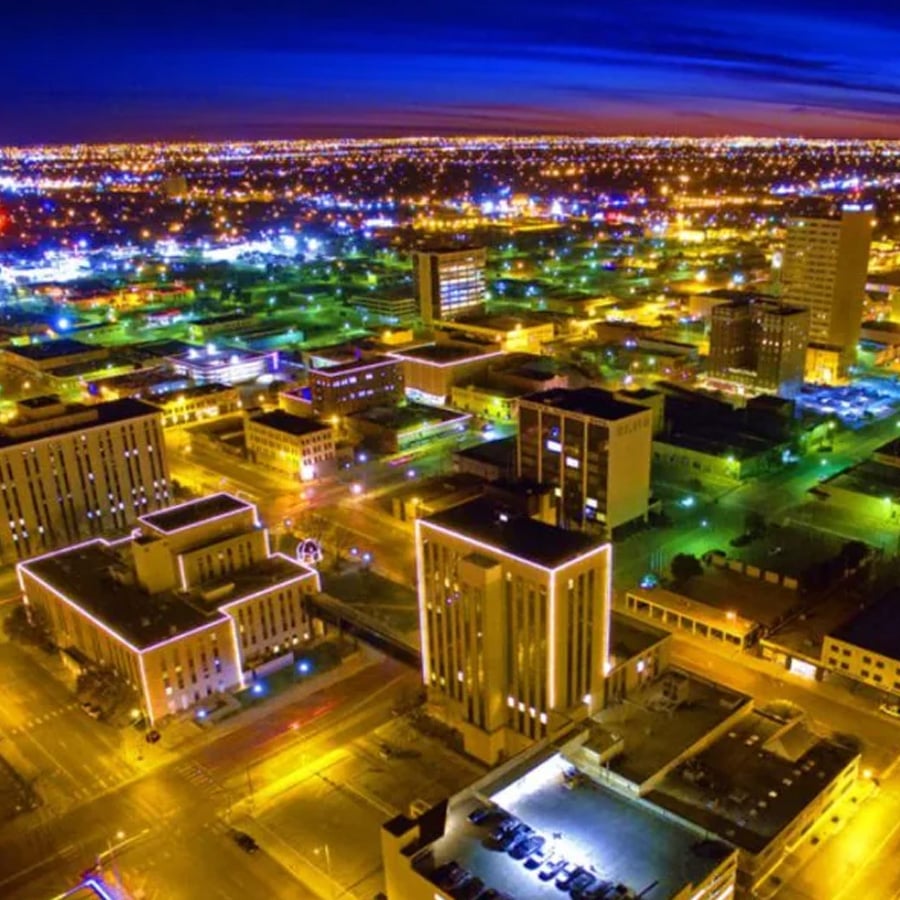 Aerial view of a city at night with brightly lit buildings, glowing streetlights, and a deep blue sky on the horizon. The city grid is clearly visible, with roads and illuminated structures.