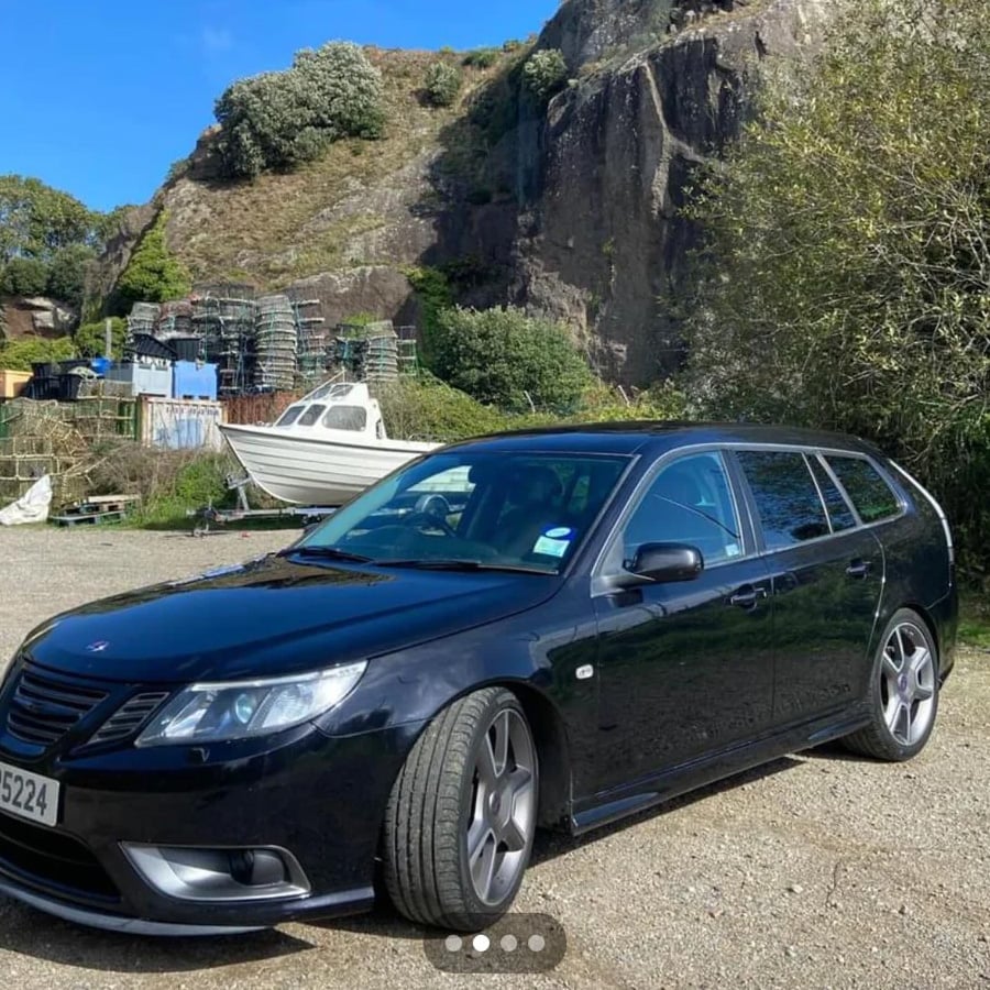 A black Saab station wagon is parked on a gravel lot near a white boat, lobster traps, and rocky cliffs with greenery in the background on a sunny day.