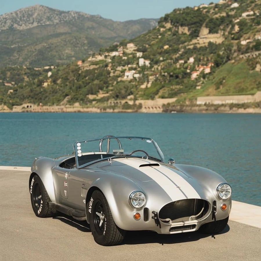 A classic silver convertible sports car with white racing stripes is parked on a waterfront road, with mountains and green hills in the background under a clear sky.
