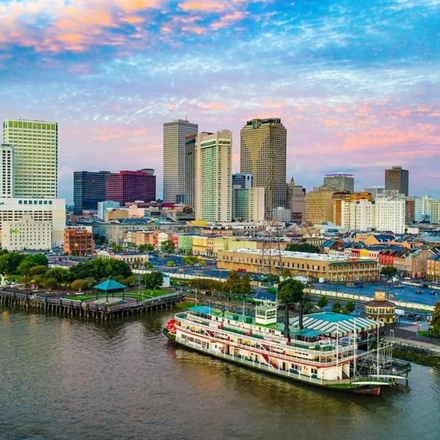 Riverboat docked by waterfront with city skyscrapers and colorful sunset sky in the background. Trees, pier, and historic-looking buildings line the shore.