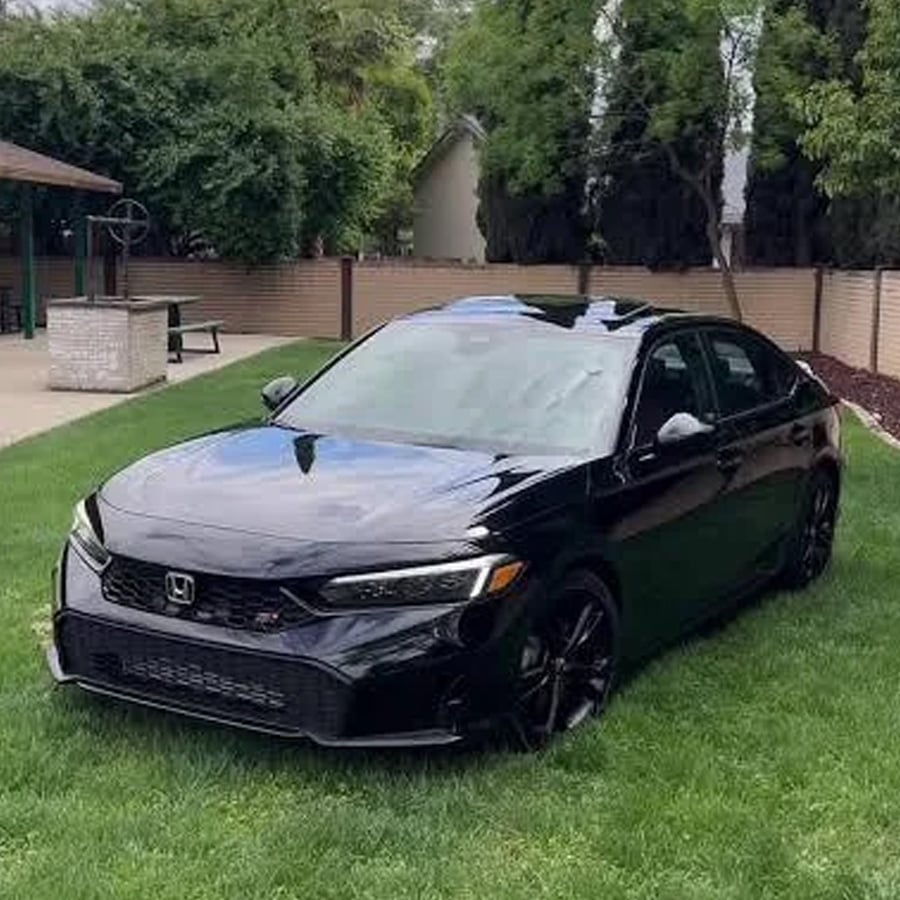A shiny black Honda sedan is parked on green grass near a picnic area, trees, and a brick wall on a partly cloudy day.