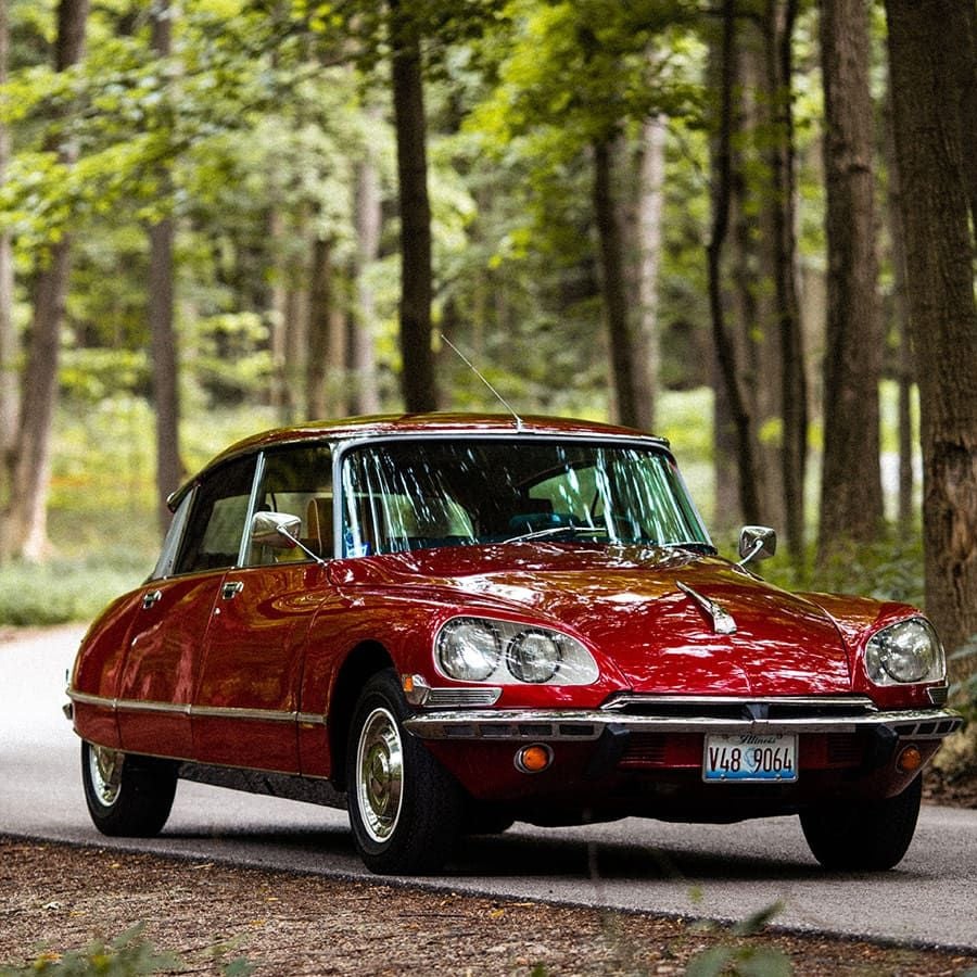 A shiny red vintage car with chrome details is parked on a road in a forested area, surrounded by tall trees and greenery.