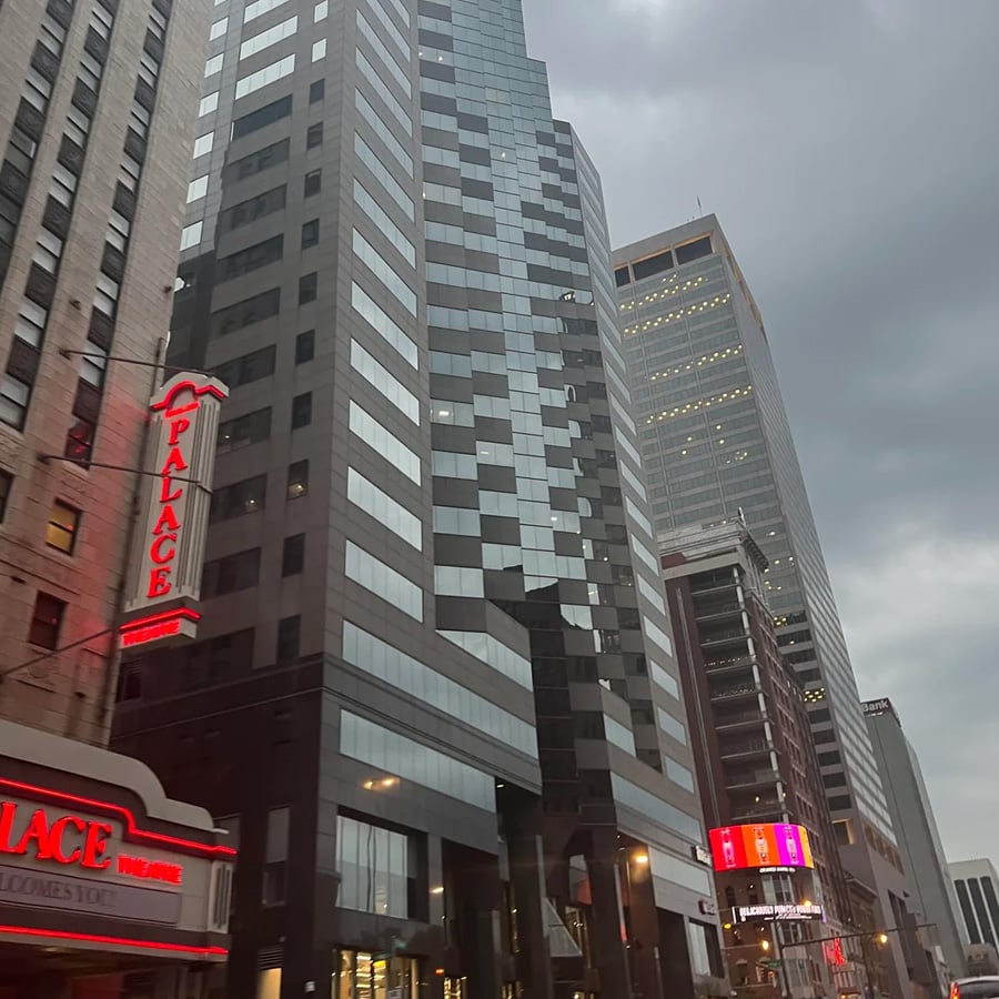 Tall modern office buildings line a city street under a cloudy sky. A lit neon sign reading “PALACE” is visible on the left side of the image, attached to a theater. Street lights and signs are also visible.
