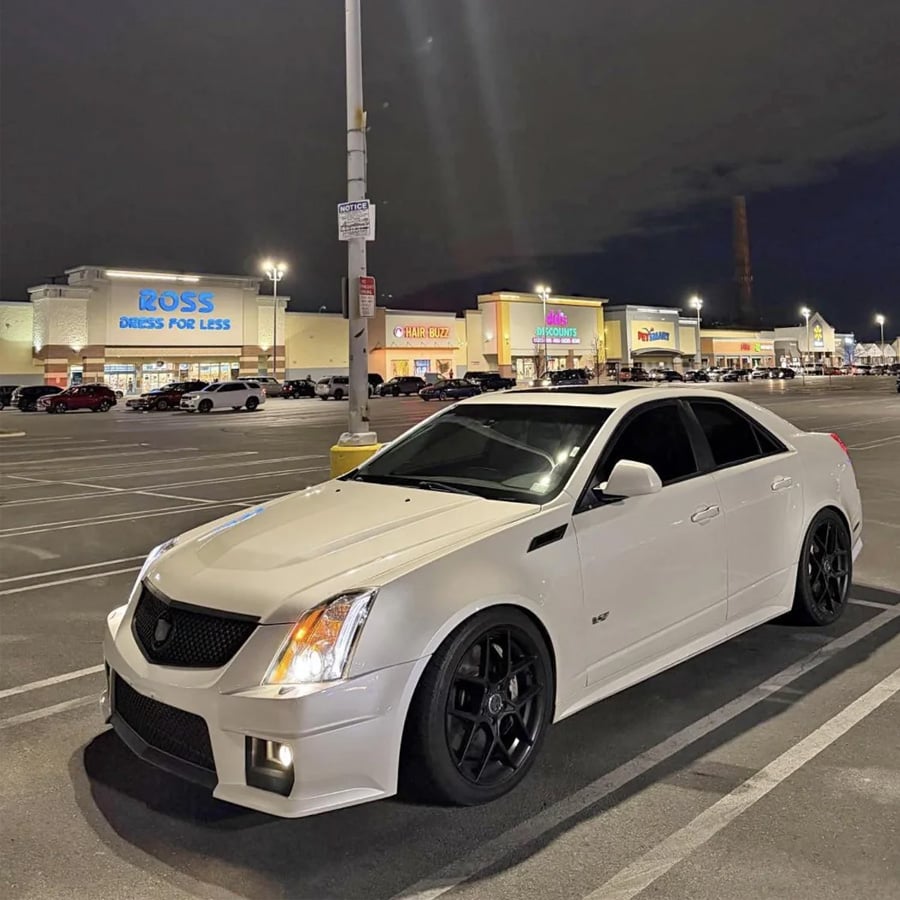 A white sports sedan with black wheels is parked in a nearly empty shopping center lot at night, with stores like Ross and PetSmart visible in the background.