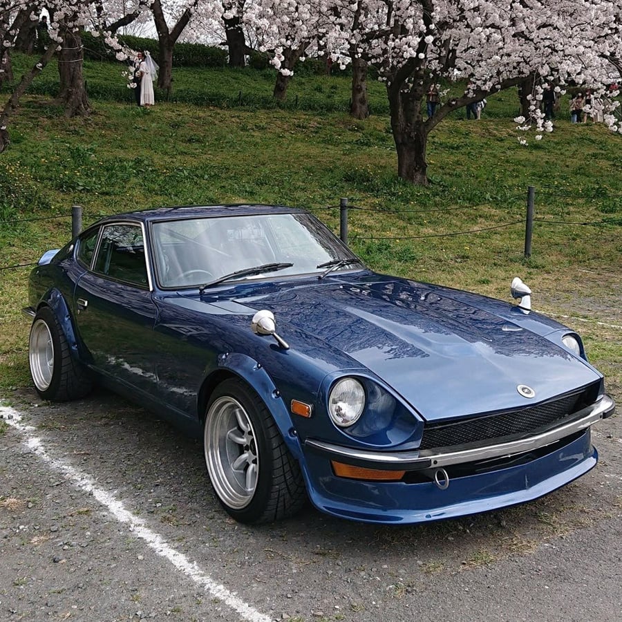 A blue vintage sports car is parked near cherry blossom trees in full bloom, with grass and blooming flowers in the background.