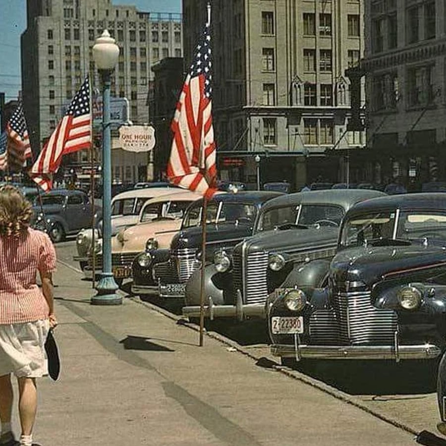 A woman walks on a city sidewalk lined with vintage cars and American flags, with tall buildings in the background.
