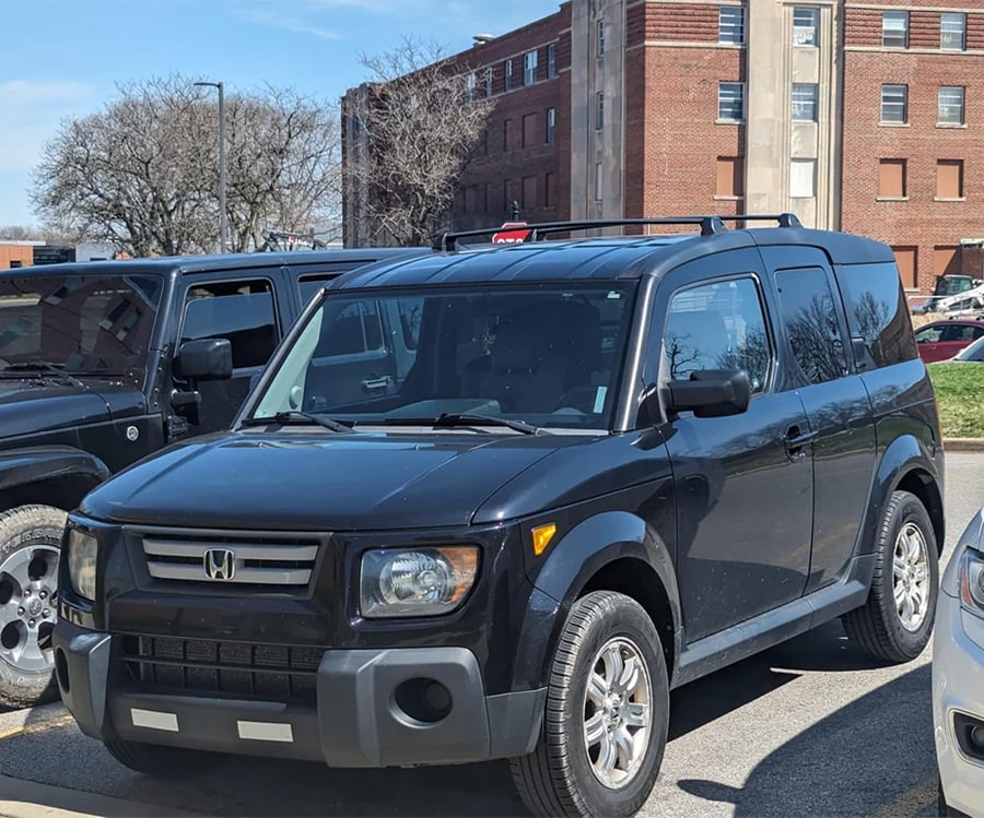 A black Honda Element SUV is parked in a parking lot, with other vehicles and a brick building in the background on a sunny day.