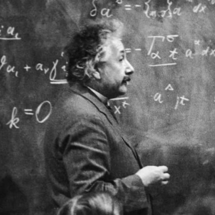 A man with curly hair and a mustache writes mathematical equations on a chalkboard while facing sideways.