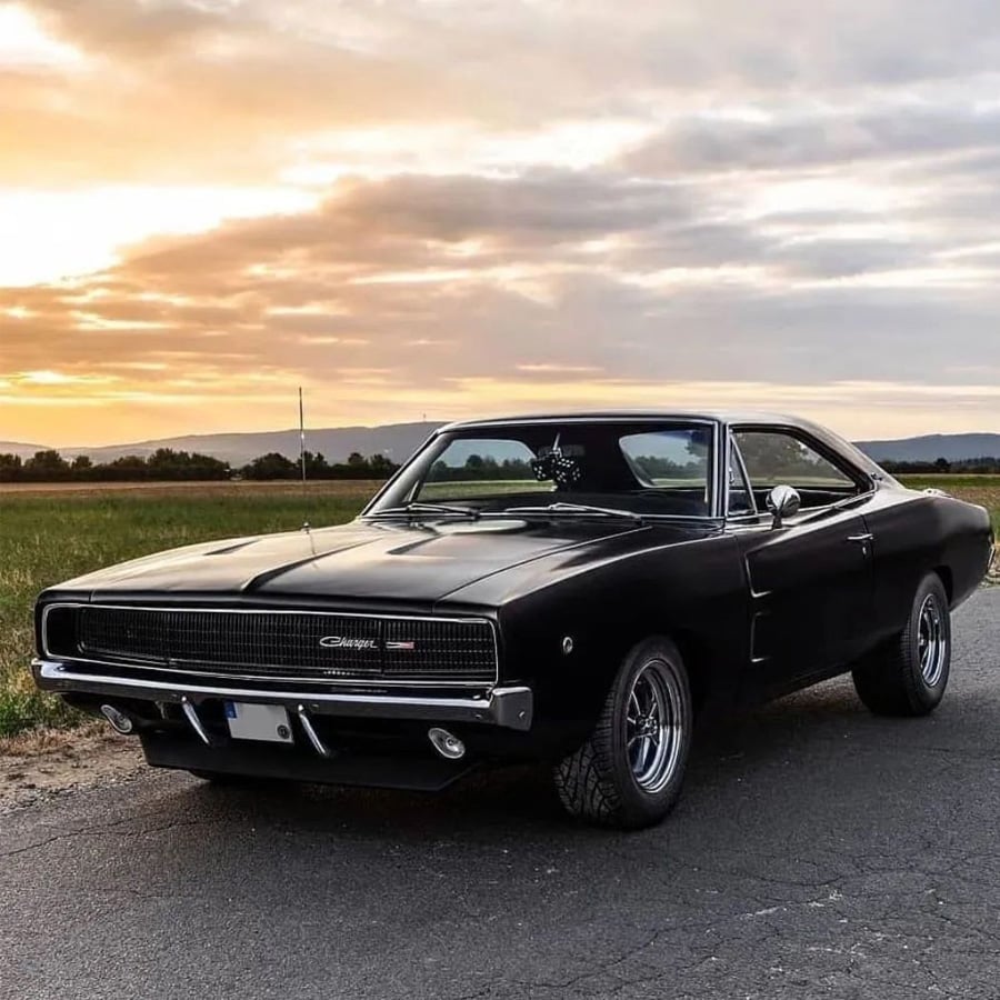 A black classic Dodge Charger muscle car is parked on a rural road at sunset, with fields and distant hills under a partly cloudy sky.