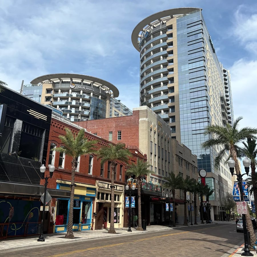 A city street lined with palm trees, historic brick buildings, and modern glass high-rises under a partly cloudy sky. The street appears quiet with no visible cars or people.