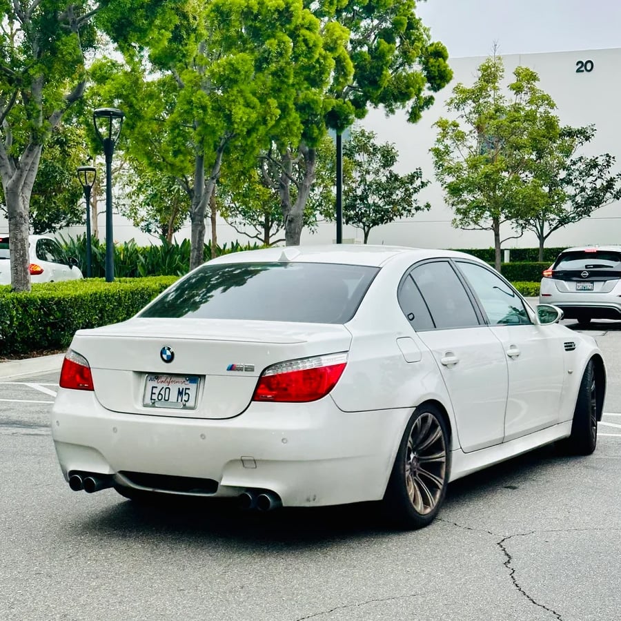 A white BMW M5 sedan is parked in a lot near green trees and bushes, with other cars and a white building in the background. The license plate reads “E60 M5.”