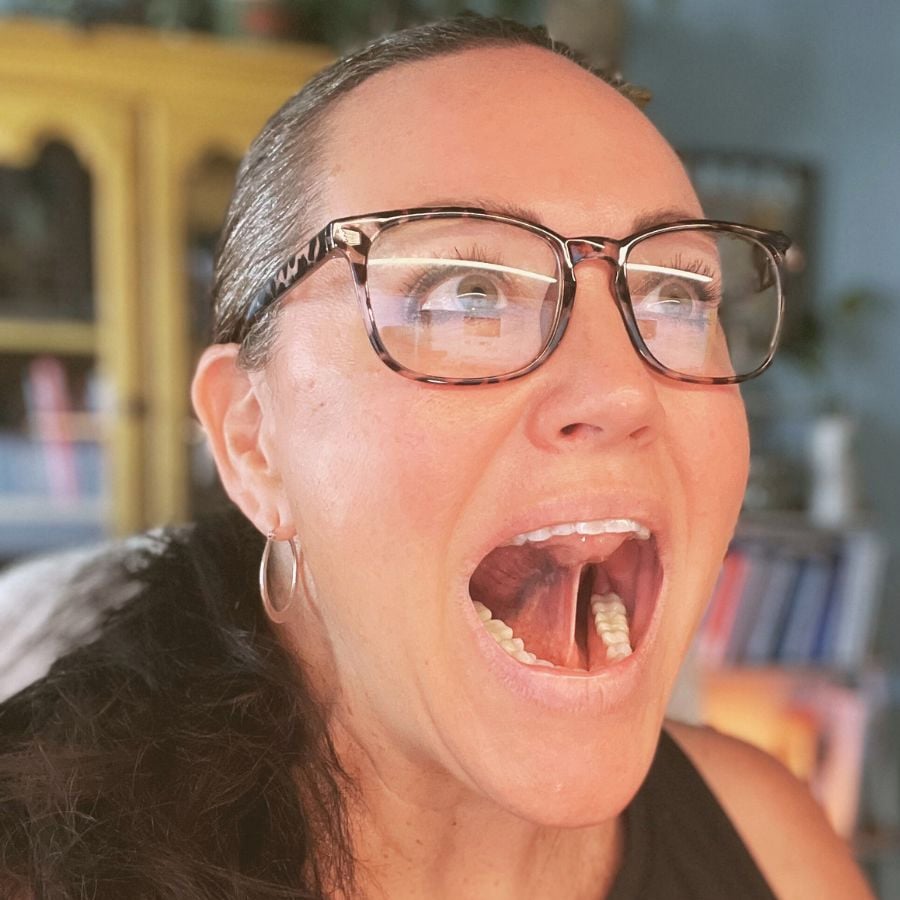 A woman with glasses and hoop earrings opens her mouth wide and looks upward, appearing surprised or excited. A bookshelf and yellow cabinet are blurred in the background.
