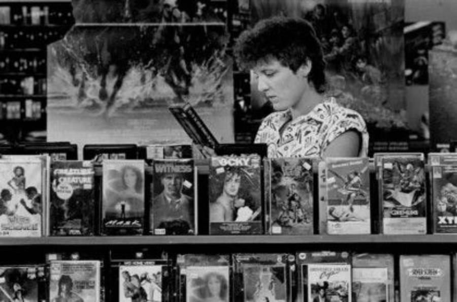 A person browses VHS tapes in a video rental store, with various movie covers visible on shelves in front. The scene appears to be from the 1980s.