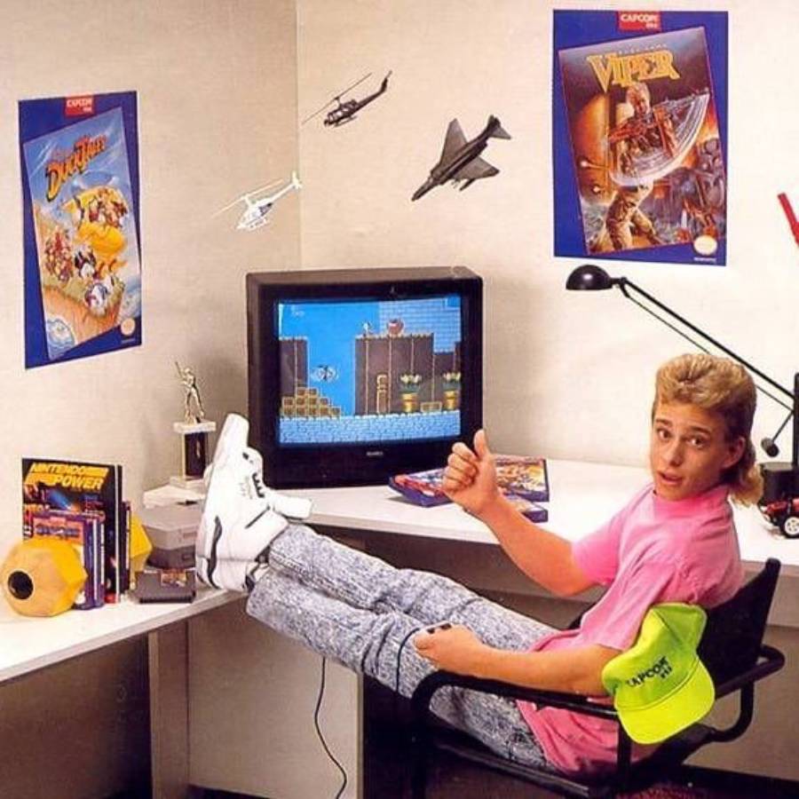 Teen boy in a pink shirt and acid-wash jeans sits at a desk, playing a retro video game. He gives a thumbs up, with posters, toy aircraft, and gaming accessories around him.