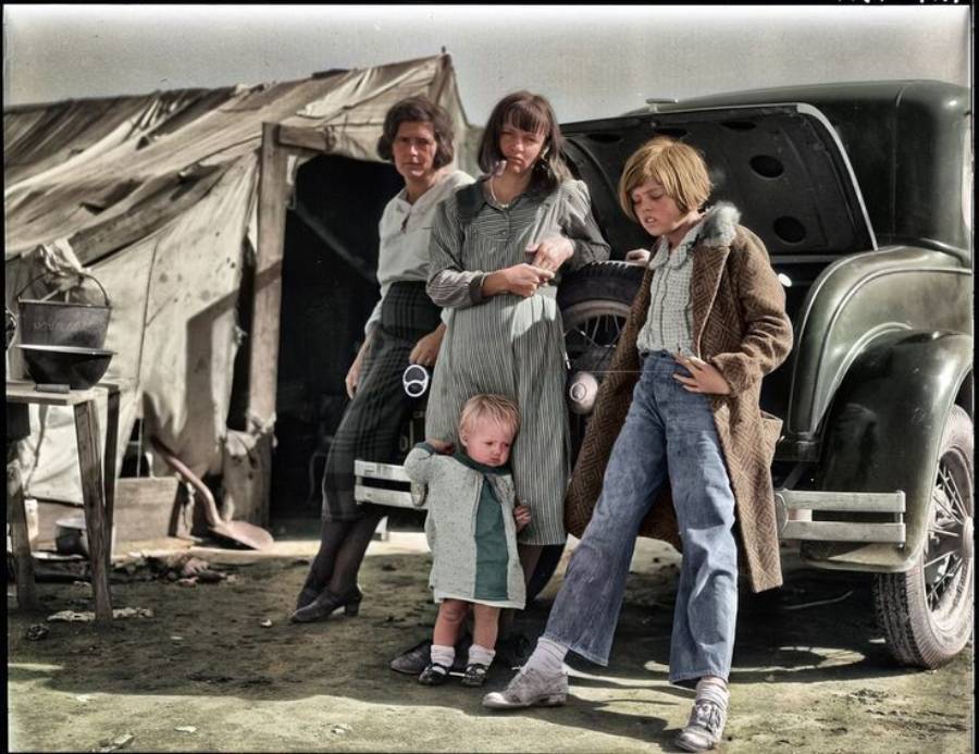 Four children stand near a car with an open trunk and a canvas tent in the background. The youngest child stands in front, while the others group together, dressed in worn clothing. The ground is dry and dusty.