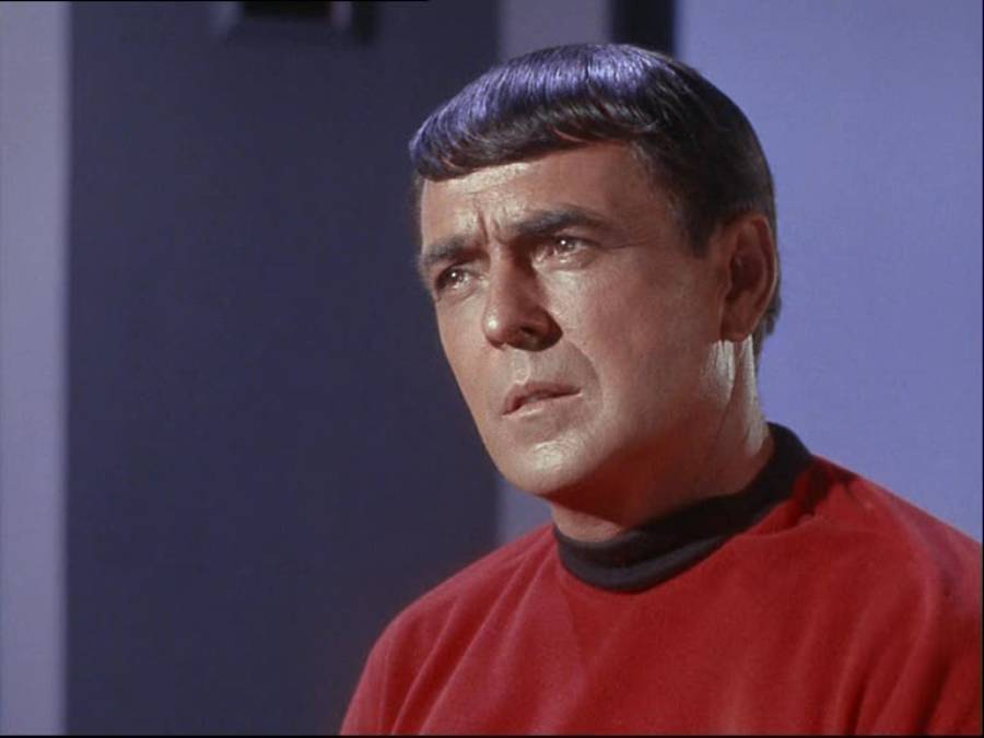 A man with short dark hair and a serious expression wears a red shirt and stands against a blurred indoor background with blue and gray tones.