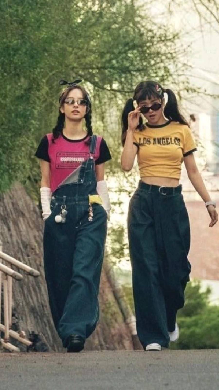 Two young women wearing wide-leg jeans, sunglasses, and colorful tops walk outdoors. One wears a pink shirt with overalls, the other a yellow “Los Angeles” shirt. Both have pigtails and playful accessories.