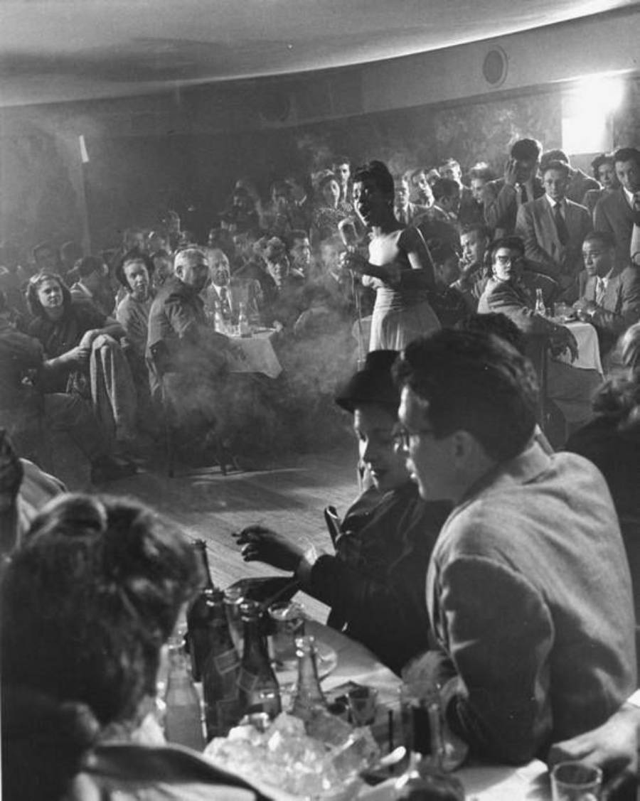 A crowded nightclub with people seated at tables, drinks on the tables, and a singer performing on stage. The room is smoky, and the audience appears captivated by the performance.