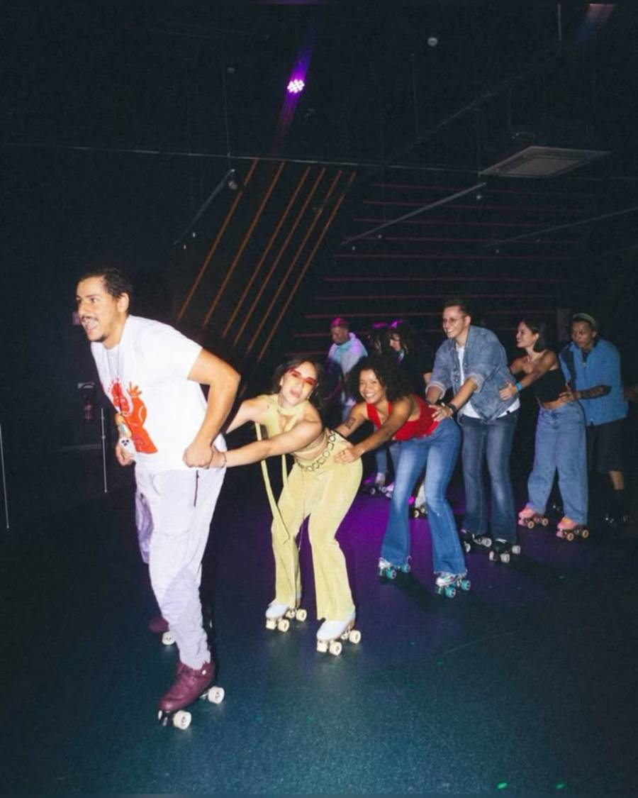 A group of people on roller skates forms a conga line under dim, colorful lighting at an indoor skating rink, smiling and having fun.