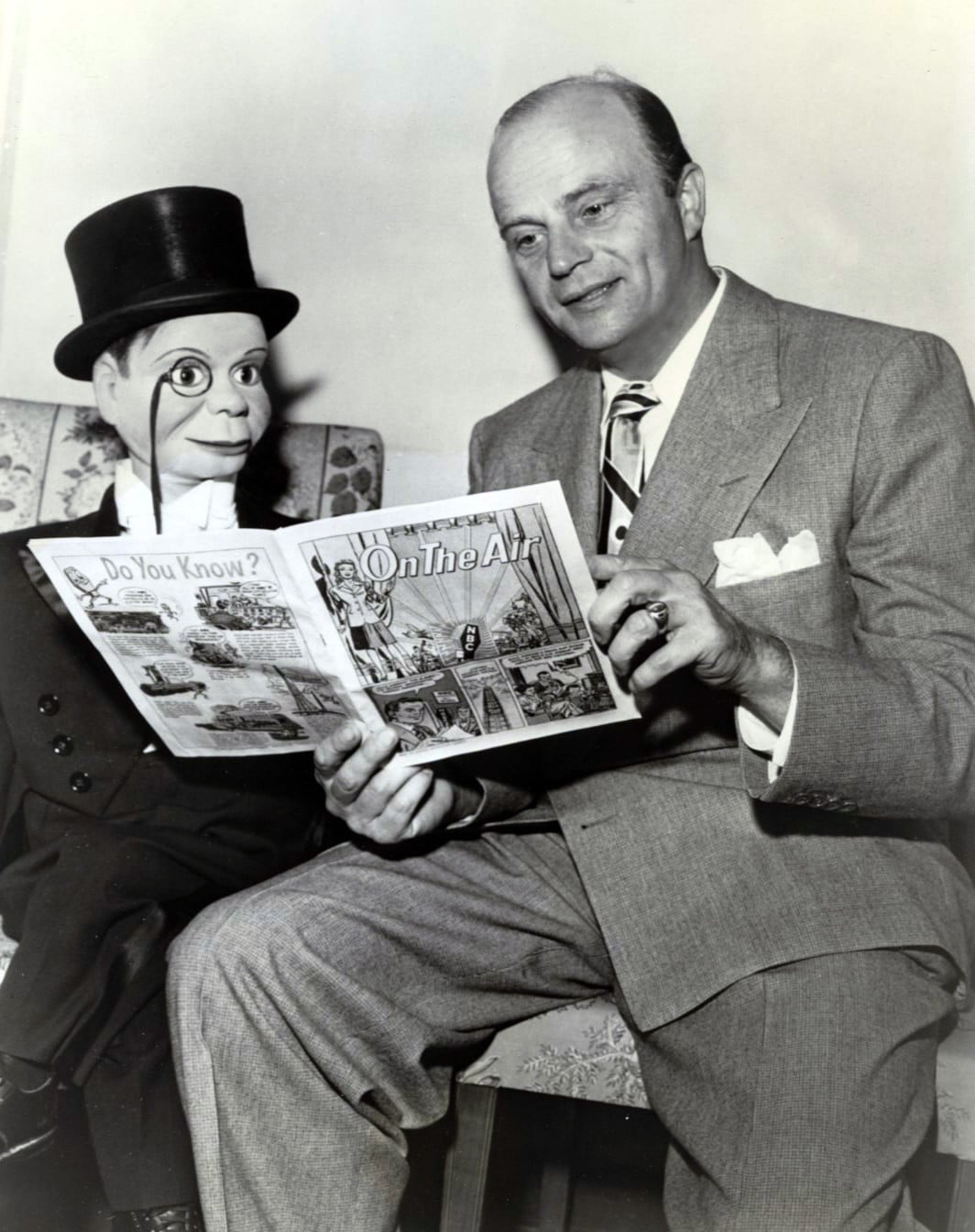 A man in a suit sits next to a ventriloquist dummy in a top hat. Both are holding and looking at an illustrated magazine titled “On The Air.”