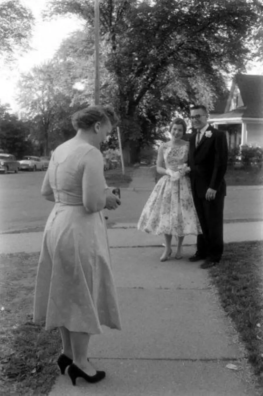 A woman in a dress stands on a sidewalk holding a camera, facing a smiling couple dressed formally and posing together near the curb in a residential neighborhood.