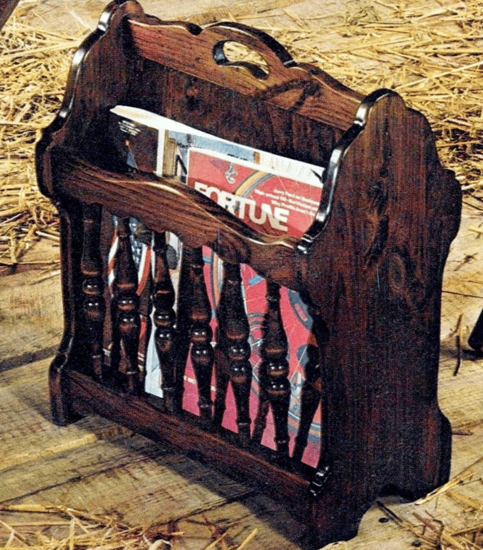 A wooden magazine rack with spindle details stands on a wooden floor, holding a red-covered magazine titled “Fortune.” Straw is scattered around the background.