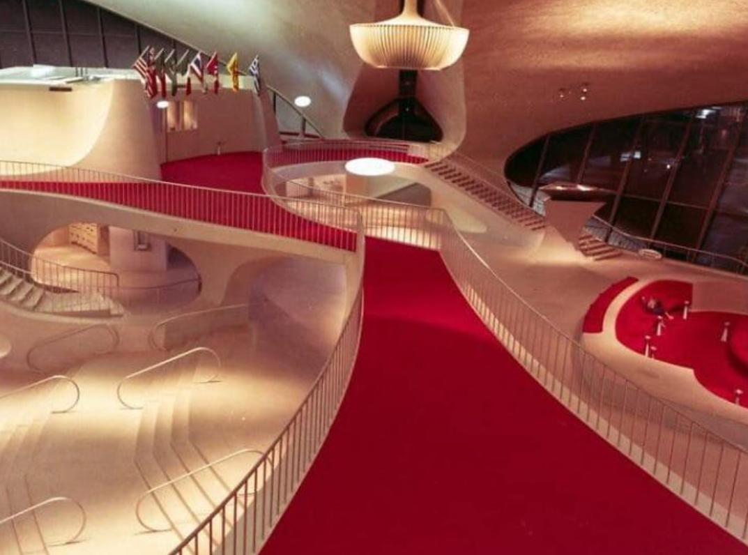 Futuristic interior with sweeping red carpet, curved white walls, large windows, modern railings, clustered flags, and hanging light fixture in a spacious, open lobby.