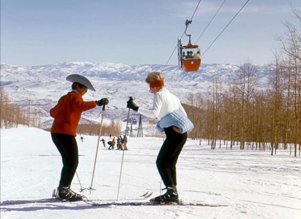Two people in winter clothing ski on a snowy slope with leafless trees nearby. A red gondola is overhead, carrying people up the mountain. Snow-covered mountains are visible in the background under a clear sky.