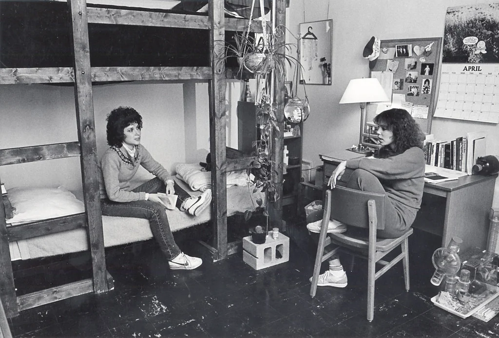 Two women sit in a dorm room with bunk beds, books, plants, and a desk. One is on a bed; the other sits on a chair. A calendar on the wall shows April. The room appears cozy and lived-in.