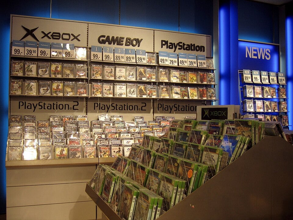 A store display shelves Xbox, PlayStation, PlayStation 2, and Game Boy games in rows, with a blue “NEWS” sign and price tags visible above the sections. Games are in plastic cases, organized neatly.
