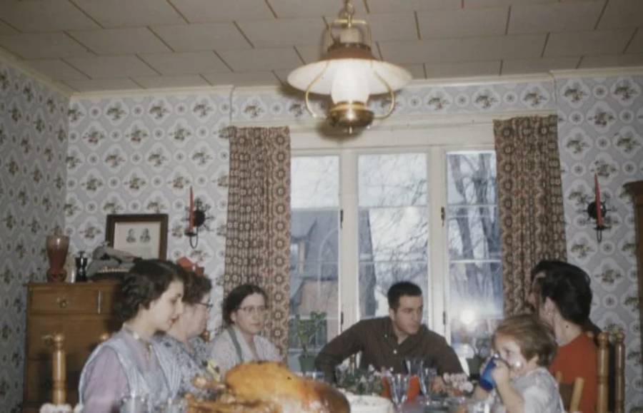 Six people sit around a dining table with a large turkey, sharing a meal in a vintage-style room with patterned wallpaper and curtains, under a hanging lamp.