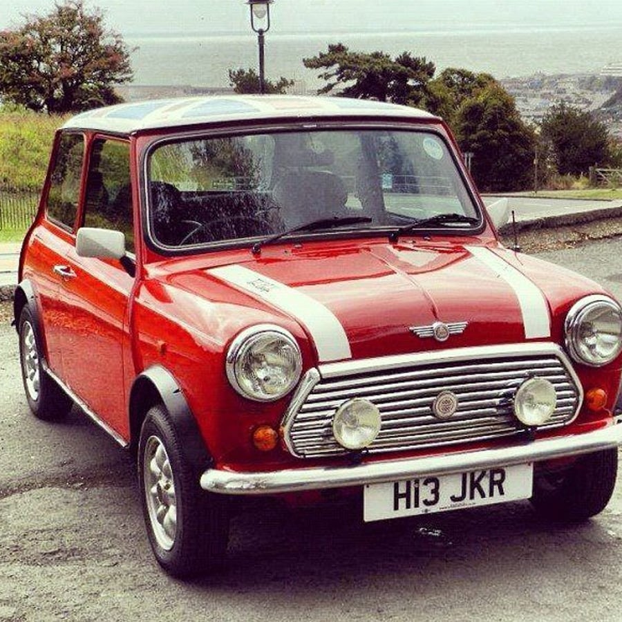 A classic red Mini Cooper with white racing stripes is parked outdoors on a road. The car has chrome bumpers and a UK license plate reading “H13 JKR.” Trees and a hilly landscape are in the background.
