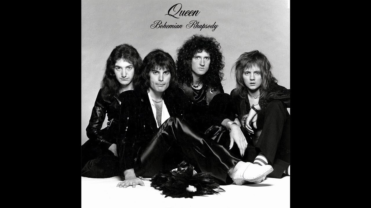 Four men sit posed together in black and white, with “Queen” and “Bohemian Rhapsody” written above them. The group is dressed in dark, stylish clothing, with a white background.