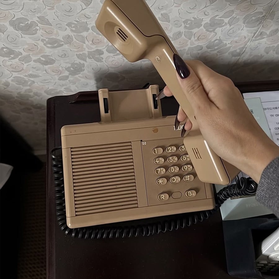 A hand with long dark nails holding the receiver of a beige retro landline telephone on a wooden table, next to a tissue box and a paper with text.