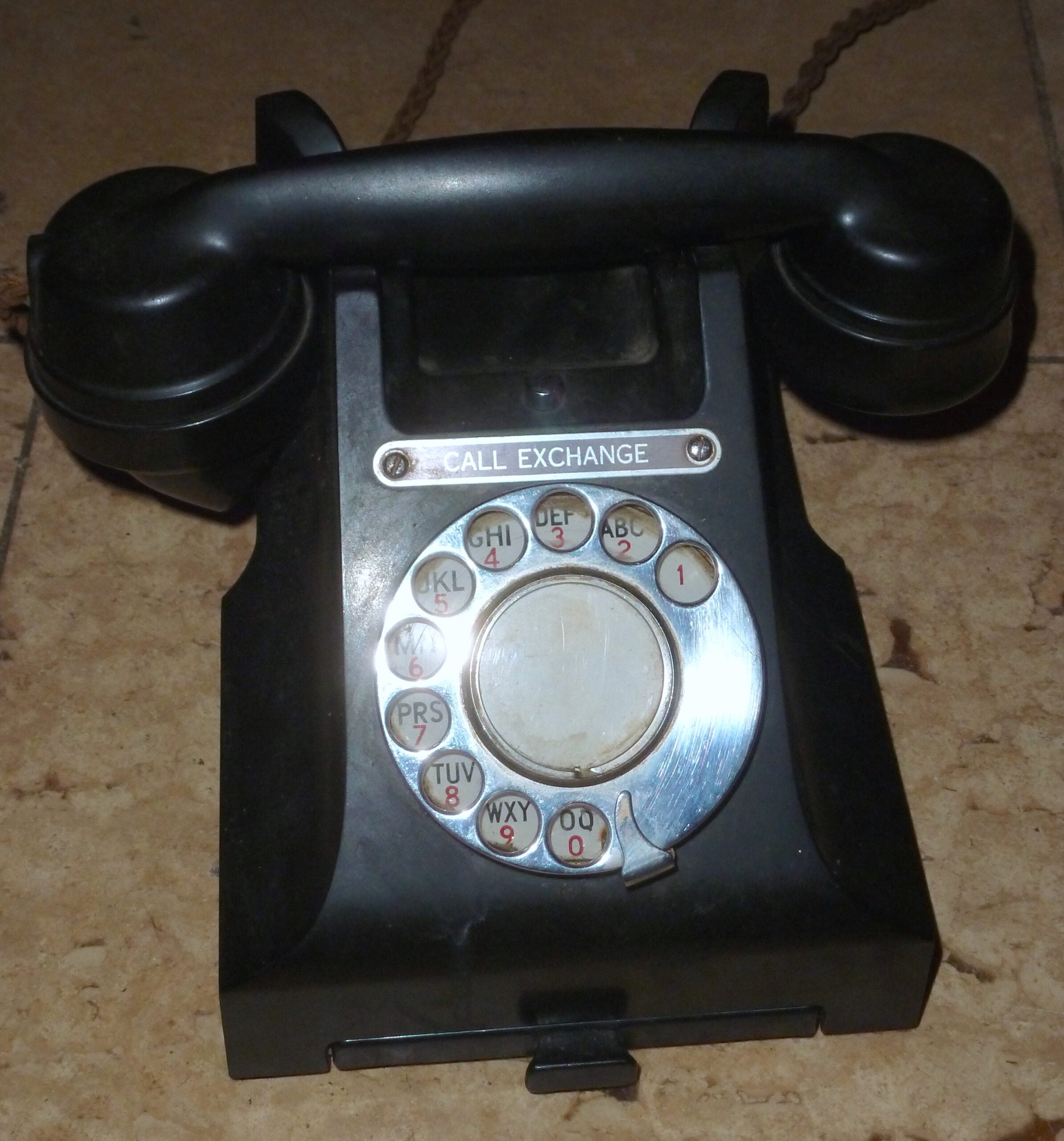 A black rotary dial telephone with a label reading “CALL EXCHANGE” sits on a brown tiled floor.
