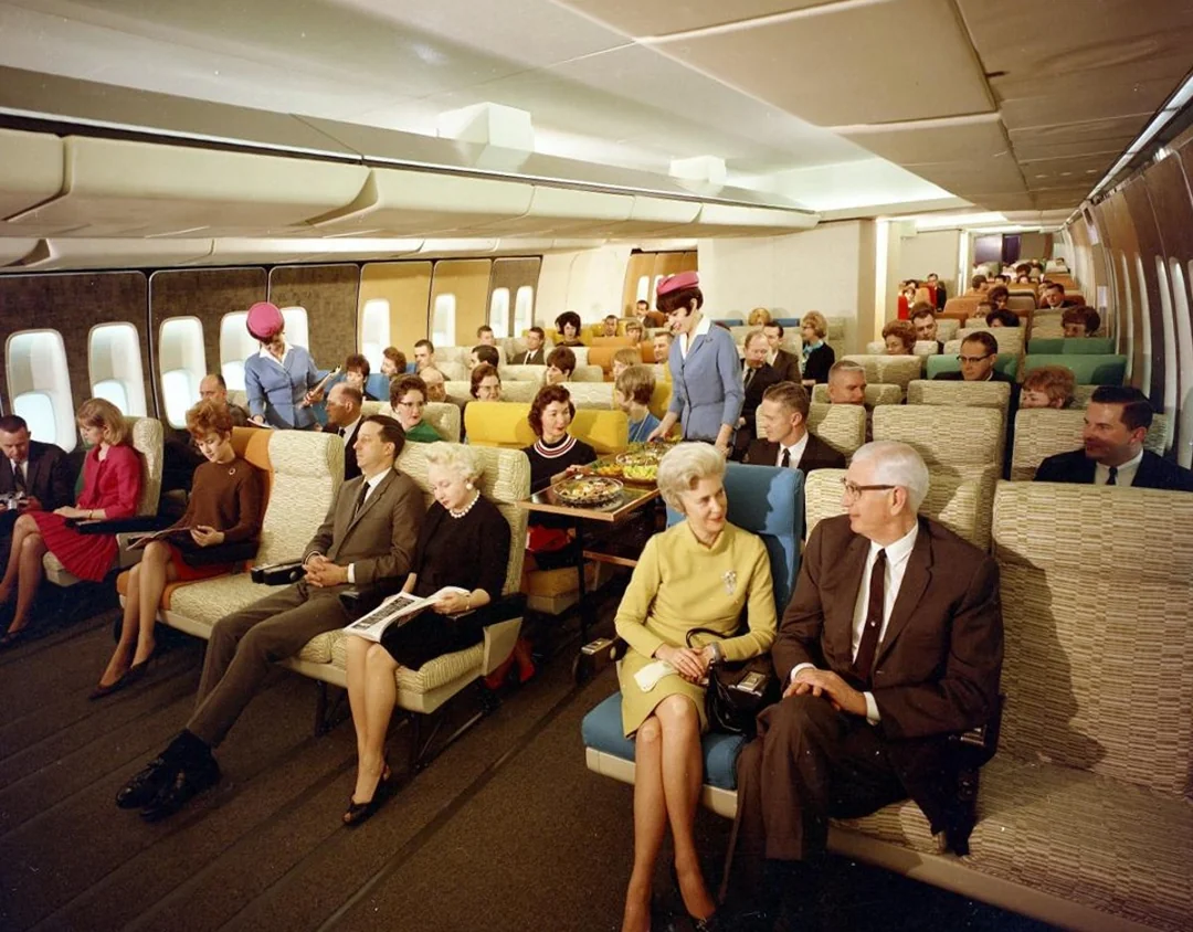 Passengers sit in colorful seats on a vintage airplane. Flight attendants in bright uniforms serve drinks, and people chat or read, creating a lively, retro in-flight scene from decades past.