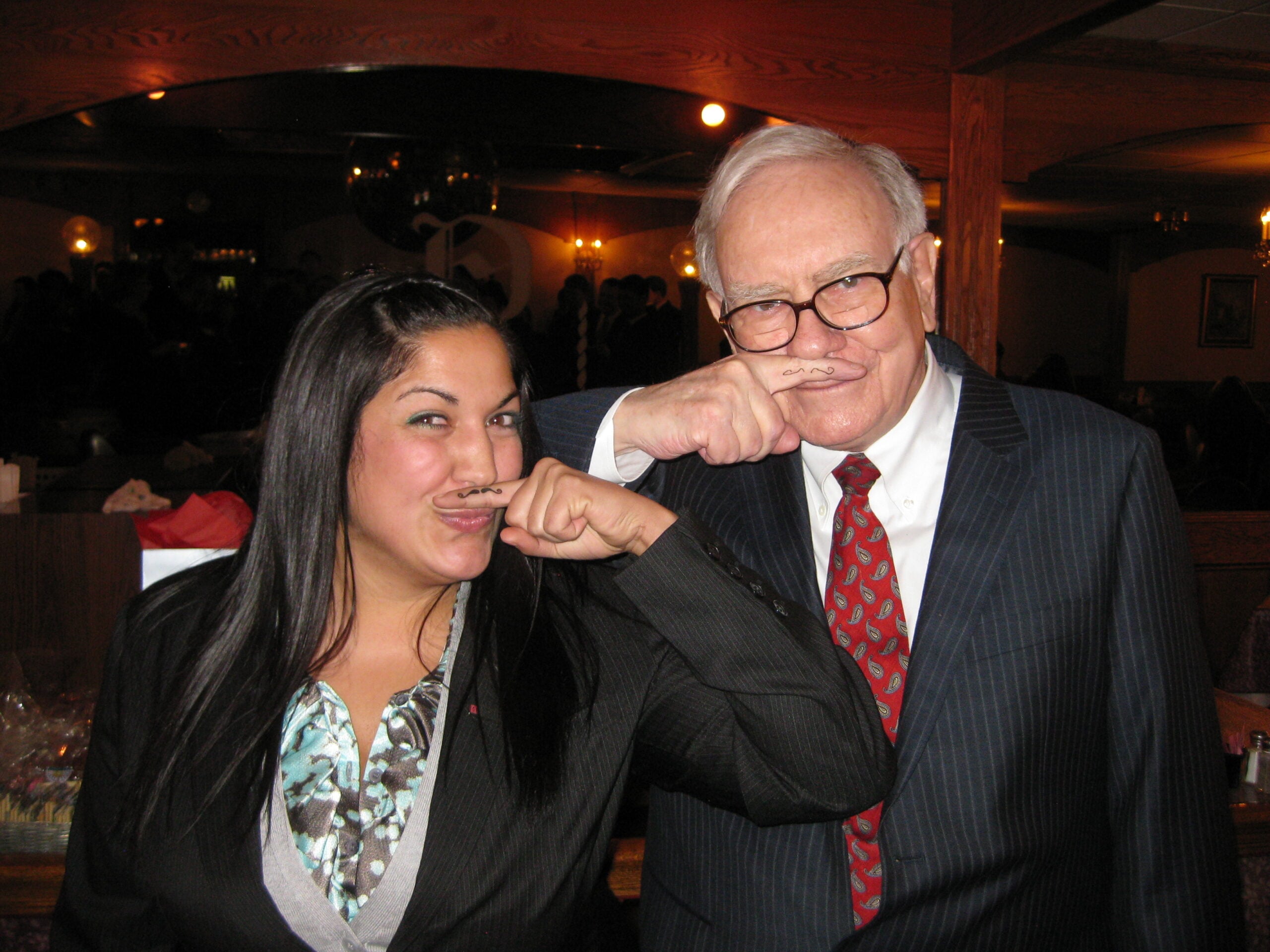 A woman and an older man in suits smile while holding their index fingers with drawn-on mustaches under their noses, pretending to have mustaches.