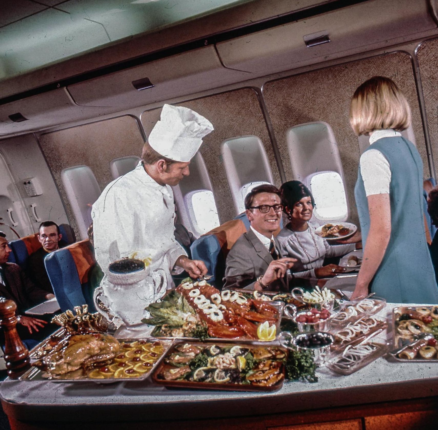 A chef and flight attendant serve gourmet dishes to well-dressed passengers on an airplane, with elaborate platters of food displayed on a cart between airplane seats.