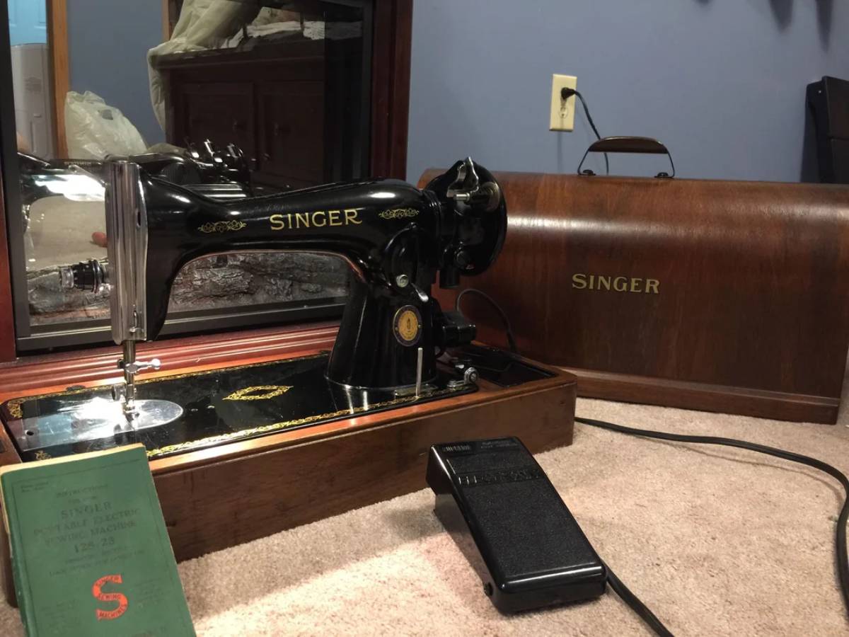 A vintage black Singer sewing machine with gold detailing sits on a carpeted floor next to its brown case, a green manual, and a black foot pedal. A mirror and cabinet are visible in the background.