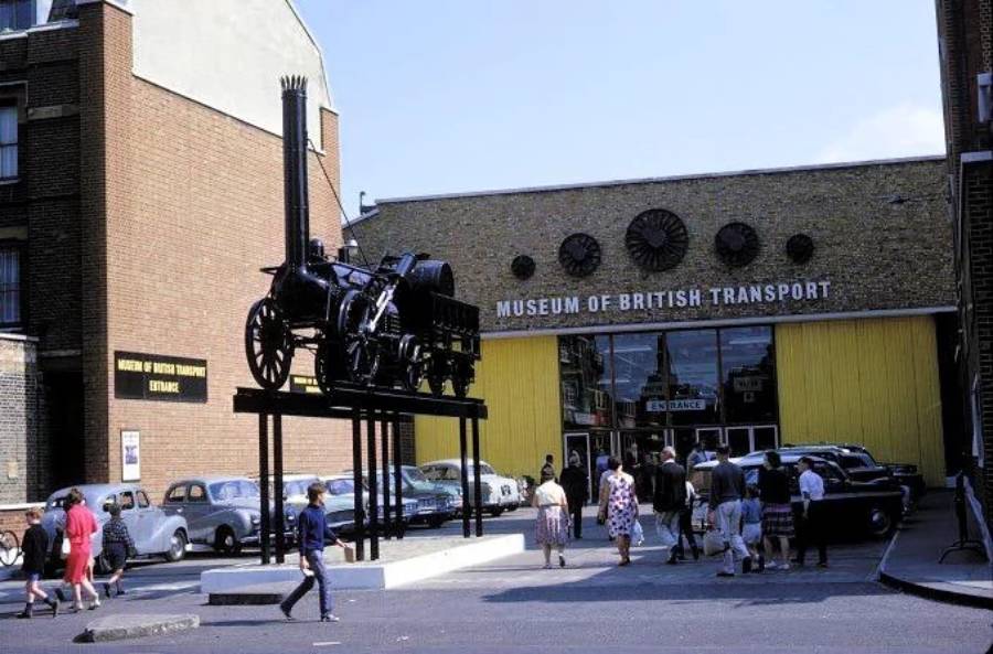 People enter the Museum of British Transport, passing vintage cars and a large black historic locomotive displayed on a tall stand outside the entrance. The building is brick with yellow doors and signage.