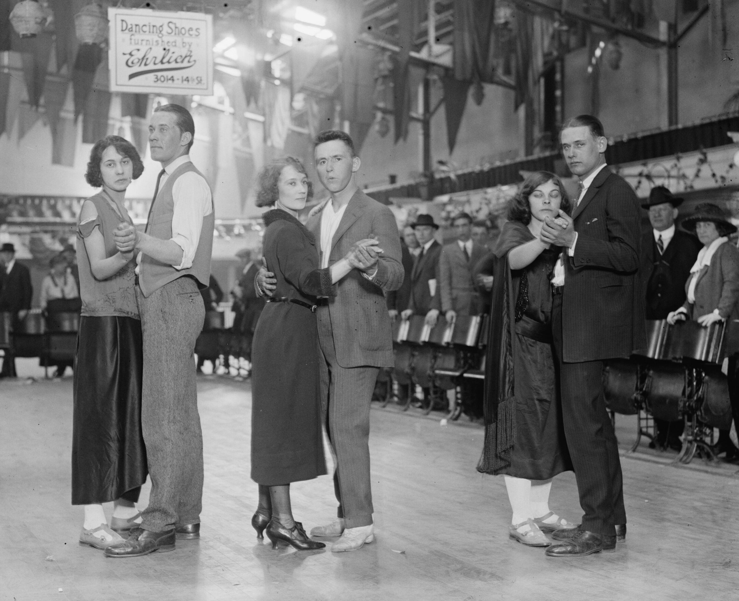 Three couples dance closely in a row on a wooden floor, with spectators and a sign advertising dancing shoes visible in the background. The scene appears to be from an earlier era, possibly the 1920s.