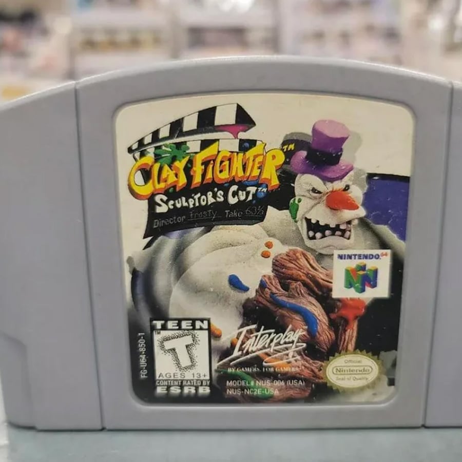 A Nintendo 64 game cartridge for “ClayFighter: Sculptor’s Cut” featuring a cartoonish angry snowman on the label. The game is rated Teen and produced by Interplay.