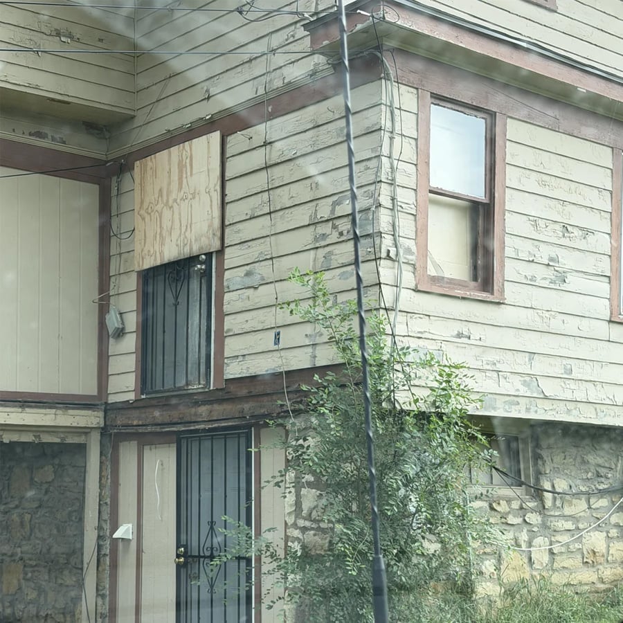 A weathered, two-story house with peeling paint, boarded-up windows, metal security doors, stone foundation, and overgrown vegetation near the front entrance.