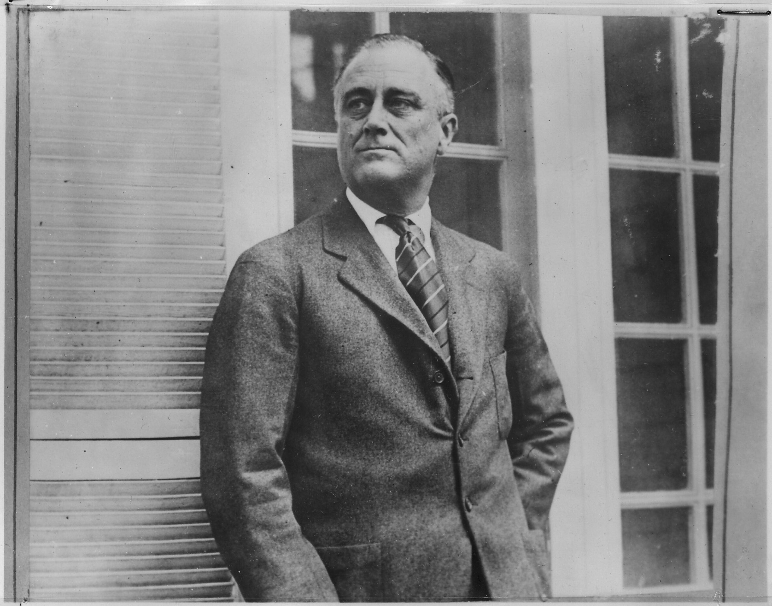 A man in a suit and striped tie stands in front of a window and shutters, looking to the side with a serious expression. The photo is black and white.