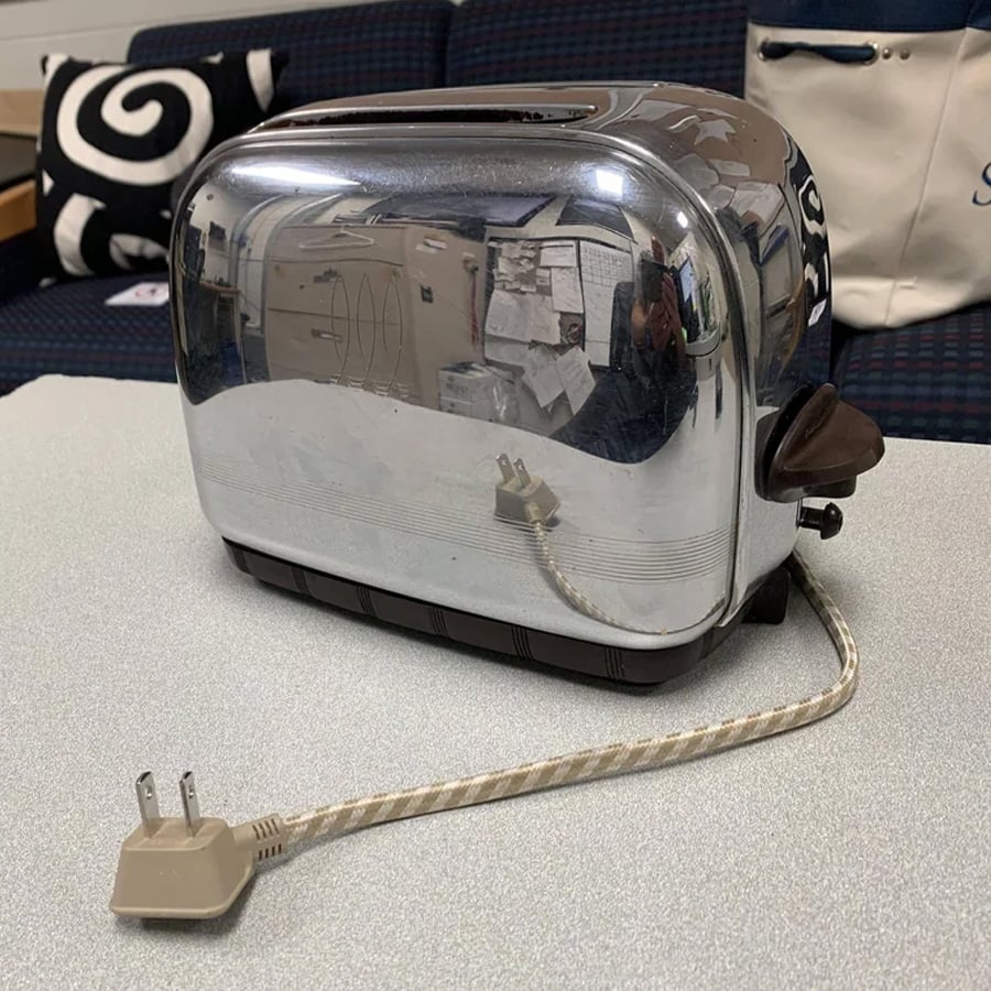 A shiny, vintage chrome toaster sits on a table, reflecting its surroundings. Its cord and plug are coiled next to it.