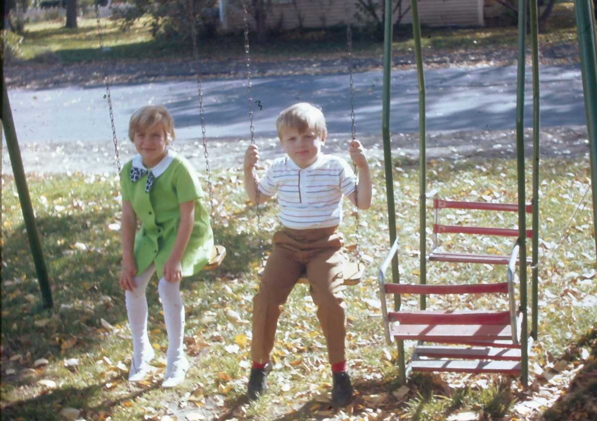 Two young children are on a swingset outdoors. The girl in a green dress sits on a swing, and the boy in striped shirt and brown pants sits on another. Fallen leaves cover the ground, and it’s a sunny day.