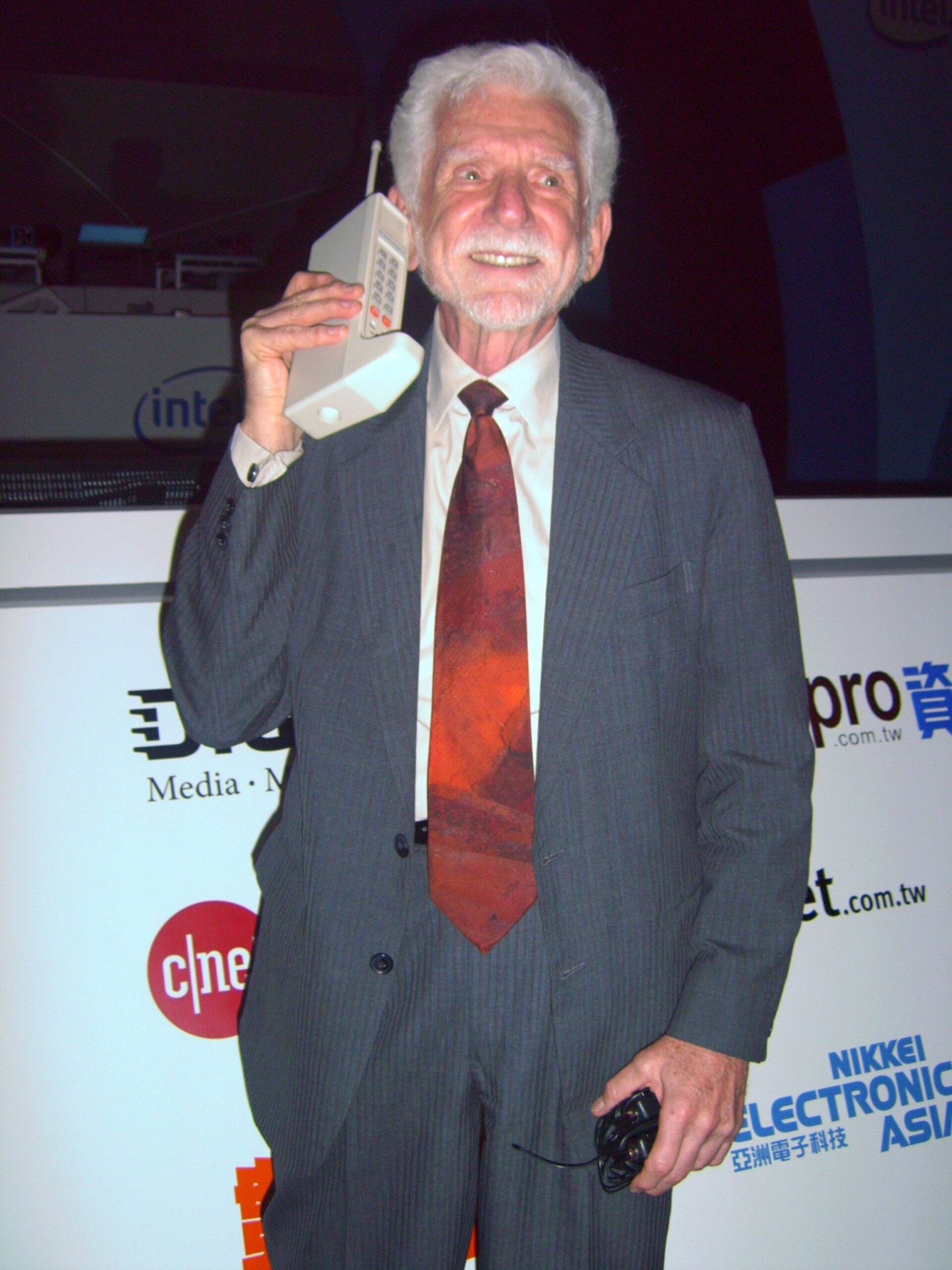 An older man in a suit smiles while holding a large, vintage mobile phone to his ear, standing in front of a backdrop with various tech company logos.
